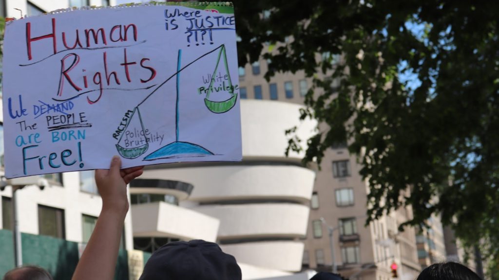 A protestor holds up a sign with a scale that shows the relationship between racism, police brutality, and white privilege. Photo by author.