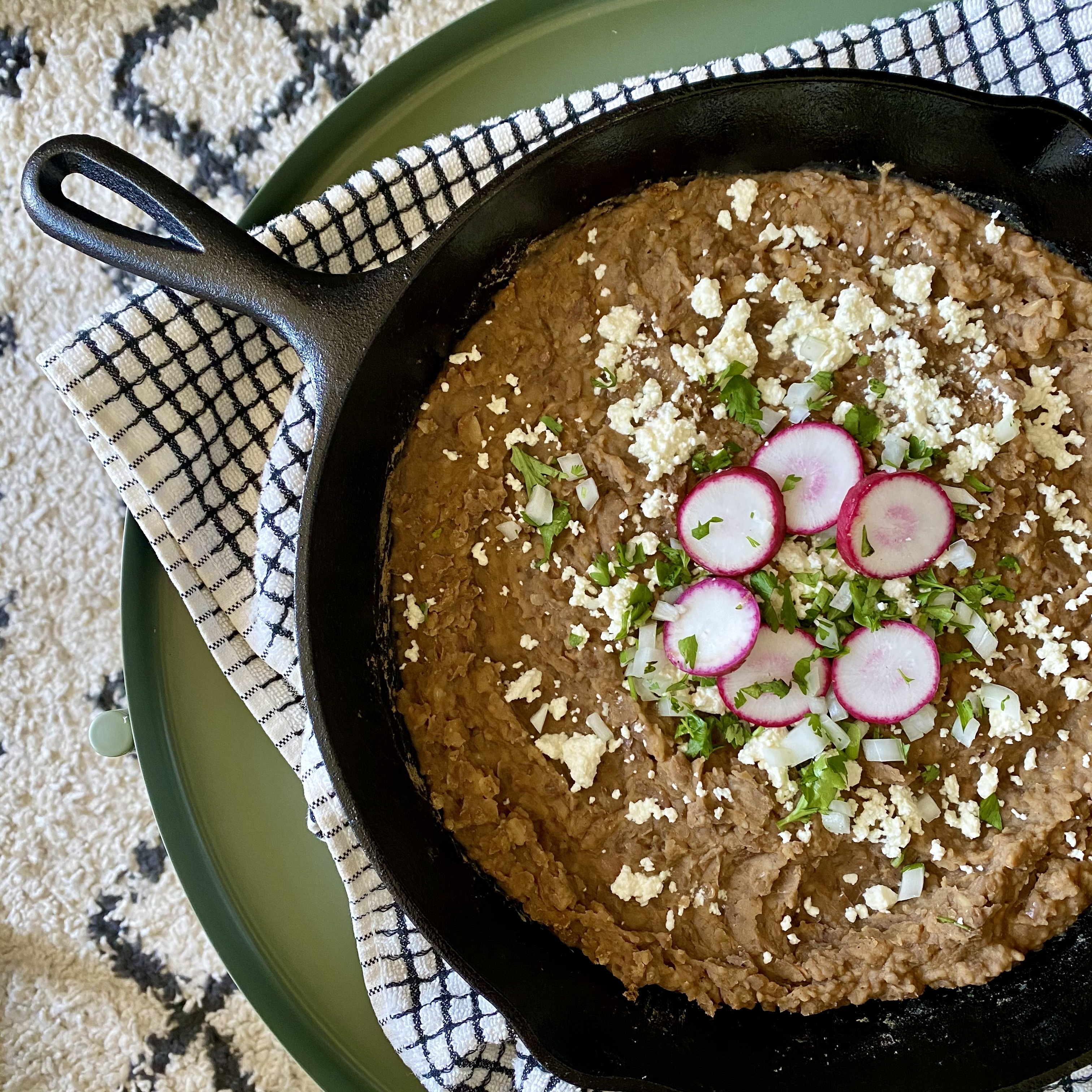 Photo of refried beans in a skillet, topped with queso fresco, radishes, and cilantro.